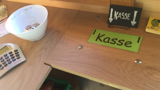 Ein Tisch mit einem Taschenrechner, einer Schale mit Wechselgeld, einem Schlitz in der Platte. Darunter ein Tresor. Ein Tisch mit einem Taschenrechner, einer Schale mit Wechselgeld, einem Schlitz in der Platte. Darunter ein Tresor.