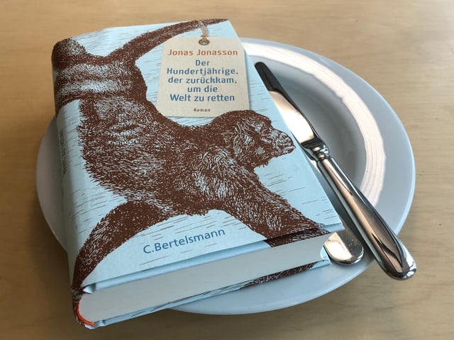 «Der Hundertjährige, der zurückkam, um die Welt zu retten» liegt auf einem weissen Teller. Messer und Gabel seitlich neben das Buch auf den Teller gelegt. «Der Hundertjährige, der zurückkam, um die Welt zu retten» liegt auf einem weissen Teller. Messer und Gabel seitlich neben das Buch auf den Teller gelegt.