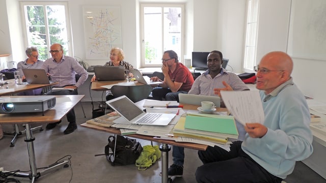 Sechs Personen sitzen an Pulten. Vor sich Computer und Papierstapel. Sechs Personen sitzen an Pulten. Vor sich Computer und Papierstapel.