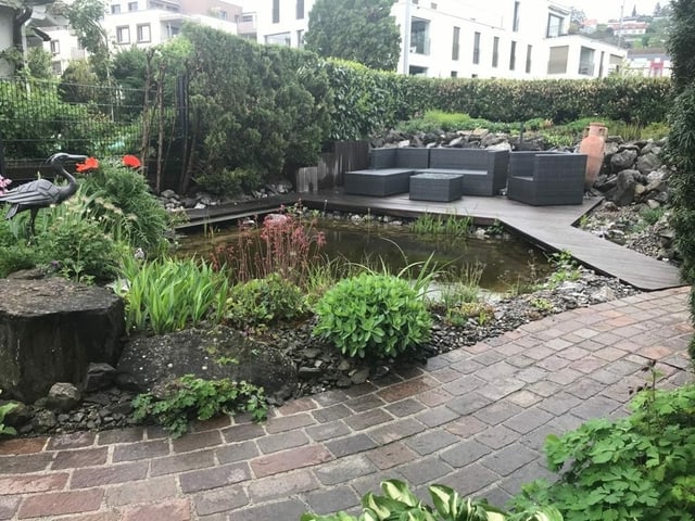 Brücke führt über ein Biotop. Dahinter sieht man eine Lounge mit Sofa und Stühlen.