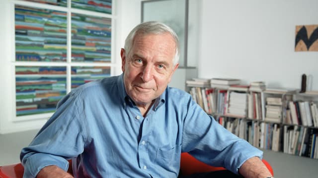 Ein Mann in blauem Hemd und mit grauen Haaren sitzt in einem Büroraum. Ein Mann in blauem Hemd und mit grauen Haaren sitzt in einem Büroraum.