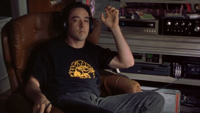 John Cusack sitzt auf einem Sessel. Er trägt Kopfhörer. John Cusack sitzt auf einem Sessel. Er trägt Kopfhörer.