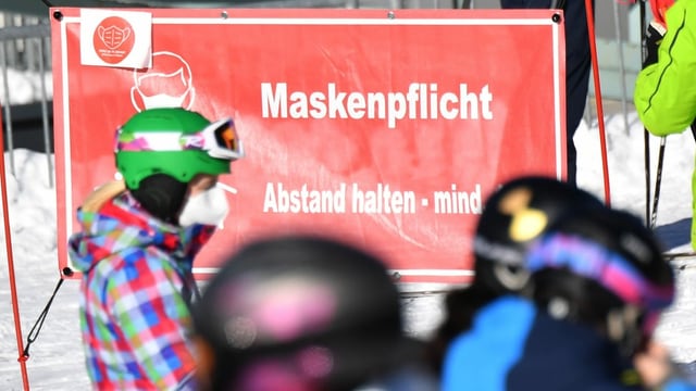 Skifahrer mit Maske vor einem Hinweisschild.