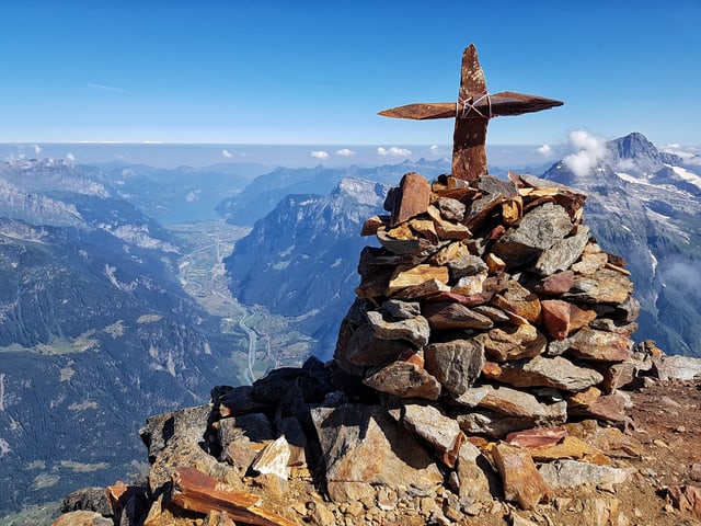 Berggipfel mit Gipfelkreuz auf Steinhaufen. Blick hinunter ins Tal und weit hinaus ins dunstige Mittelland.