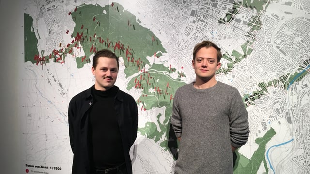 Zwei junge Männer mit kurzen Haaren und schwarzem und grauem Pullover stehen vor einem grossen Stadtplan von Zürich. Zwei junge Männer mit kurzen Haaren und schwarzem und grauem Pullover stehen vor einem grossen Stadtplan von Zürich.