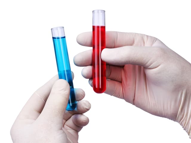 Zwei Hände halten je ein Reagenzglas mit einer blauen und einer roten Flüssigkeit