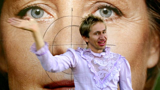 Mann in Bluse und mit Lippenstift vor einem Plakat von Angela Merkel. Mann in Bluse und mit Lippenstift vor einem Plakat von Angela Merkel.