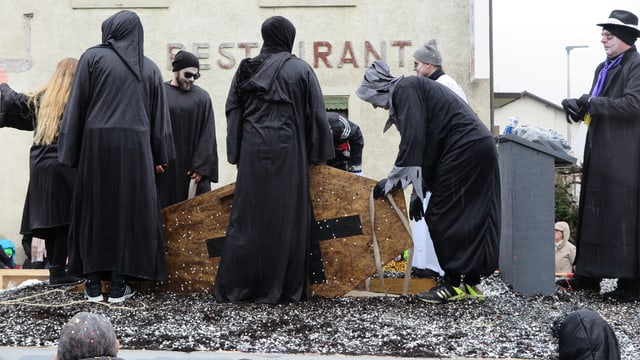 Schwarz gekleidete Figuren versenken einen Sarg in einem Boden aus Konfetti Schwarz gekleidete Figuren versenken einen Sarg in einem Boden aus Konfetti