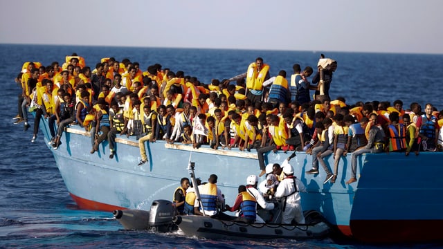 Ein Schlauchboot mit Soldaten vor einem überfüllten Holzboot. Ein Schlauchboot mit Soldaten vor einem überfüllten Holzboot.