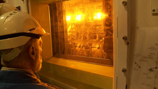 Ein Mann mit Schutzhelm blickt durch ein Fenster auf ein hell beleuchtetes Gerät Ein Mann mit Schutzhelm blickt durch ein Fenster auf ein hell beleuchtetes Gerät
