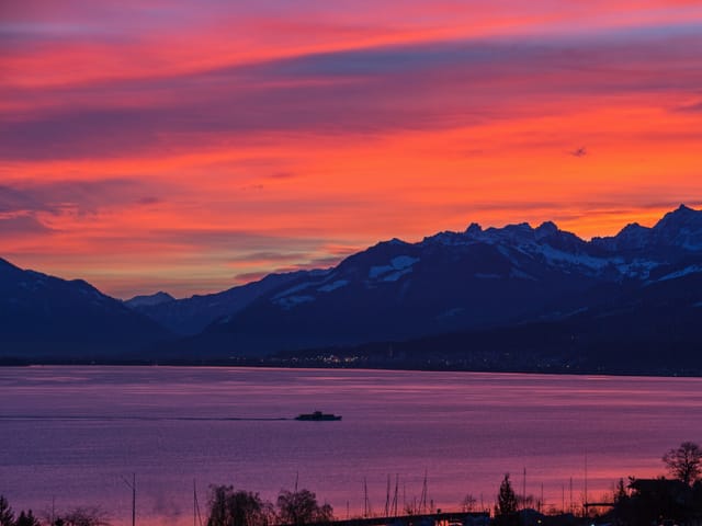 Das Morgenrot spiegelt sich im See. Dazwischen liegt eine dunkle Bergkette. Auf dem See befindet sich ein Schiff.