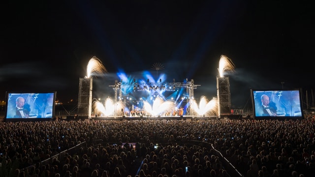 Blick von weitem auf eine grosse Bühne mit einem Orchester. Auf der Bühne ist Feuerwerk zu sehen. Blick von weitem auf eine grosse Bühne mit einem Orchester. Auf der Bühne ist Feuerwerk zu sehen.