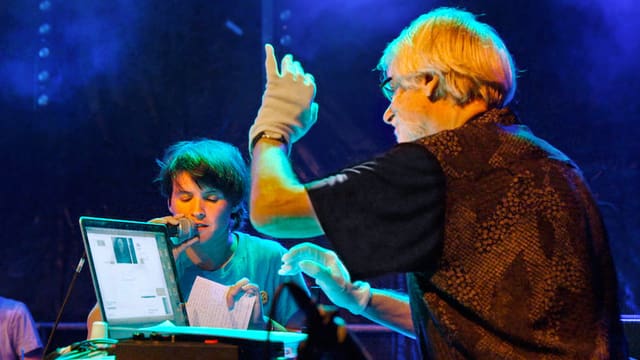 Ein Mann sitzt vor einem Laptop und hat die linke Hand angehoben, ein Junge singt in ein Mikrofon. Ein Mann sitzt vor einem Laptop und hat die linke Hand angehoben, ein Junge singt in ein Mikrofon.