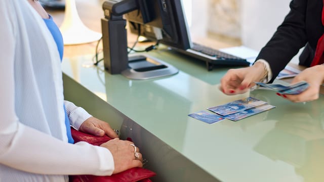 Frau steht am Bankschalter, Angestellte zählt 100er-Noten auf den Tresen. Frau steht am Bankschalter, Angestellte zählt 100er-Noten auf den Tresen.