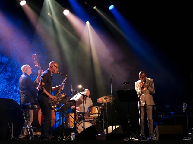 Eine Momentaufnahme während eines Jazzkonzerts. Die dunkle Bühne ist mit einzelnen, farbigen Scheinwerfern beleuchtet. Die Mitglieder der Band «Still Dreaming» mit Kontrabass, Schlagzeug, Trompete und Saxophon stehen dicht beieinander und spielen ihre Instrumente. Sie schauen einander nicht an, und bilden doch eine Einheit. Eine Momentaufnahme während eines Jazzkonzerts. Die dunkle Bühne ist mit einzelnen, farbigen Scheinwerfern beleuchtet. Die Mitglieder der Band «Still Dreaming» mit Kontrabass, Schlagzeug, Trompete und Saxophon stehen dicht beieinander und spielen ihre Instrumente. Sie schauen einander nicht an, und bilden doch eine Einheit.