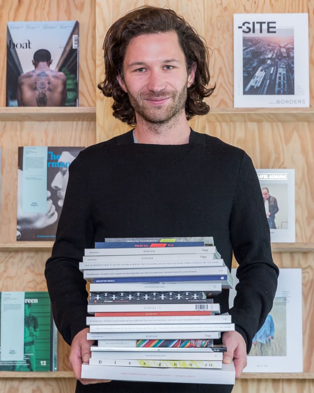 Ein junger Mann posiert mit einem Stapel Zeitschriften in den Händen vor einem Bücherregal. Ein junger Mann posiert mit einem Stapel Zeitschriften in den Händen vor einem Bücherregal.