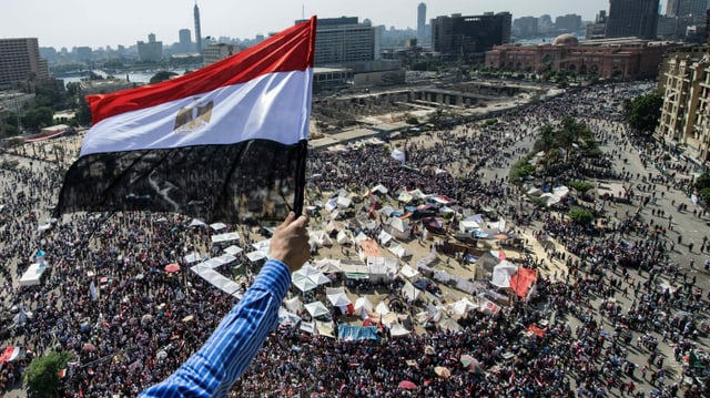 Blick auf den Platz, der voll mit Menschen ist. Im Vordergrund ein Arm, der eine ägyptische Flagge hält. Blick auf den Platz, der voll mit Menschen ist. Im Vordergrund ein Arm, der eine ägyptische Flagge hält.