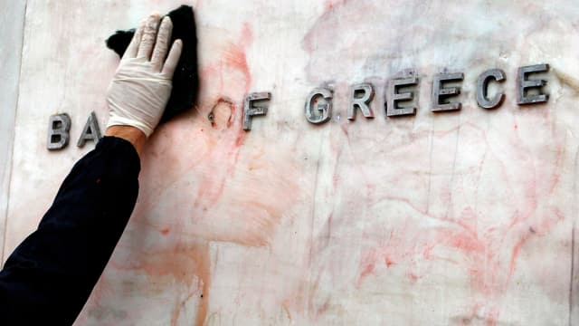 Eine Hand wischt das Logo der Bank of Greece ab. 