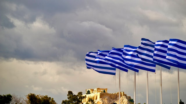 Griechische Flaggen im Wind, im Hintergrund ist die Akropolis erkennbar, grauer Himmel.