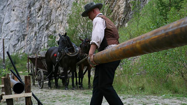 Ein Mann trägt ein Rohr, um eine Pipeline zu bauen. Im Hintergrund stehen vor einer Felswand zwei Pferde mit einem Wagen. Ein Mann trägt ein Rohr, um eine Pipeline zu bauen. Im Hintergrund stehen vor einer Felswand zwei Pferde mit einem Wagen.