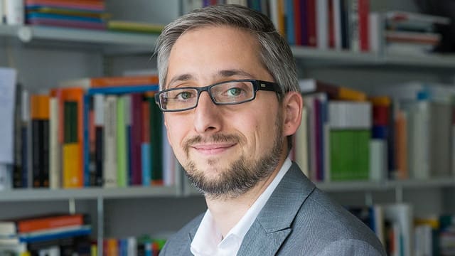 Ein Mann mit grauem Haar und Bart vor einem Büchergestell. Ein Mann mit grauem Haar und Bart vor einem Büchergestell.