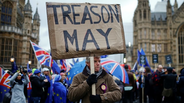 The man demonstrates in front of the Parliament, behind him with umbrellas and English flags.