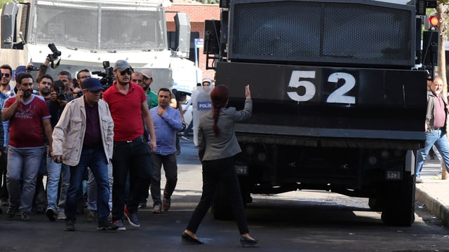 Frau hält ein Armeefahrzeug auf, daneben eine Gruppe wütender Männer. 