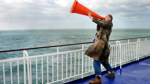 Ein steht an der Reling eines Schiffs und hält sich einen Leitkegel vor den Mund. Ein steht an der Reling eines Schiffs und hält sich einen Leitkegel vor den Mund.