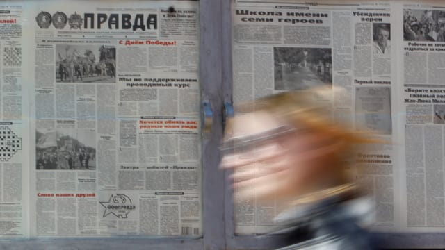 Eine Frau (verschwommen) geht an einem Schaukasten mit aufgehängten Zeitungsseiten vorbei.