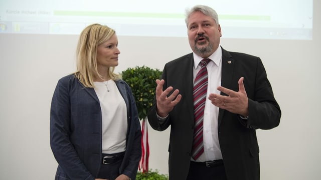 Eine Frau steht neben einem Mann in einem Verwaltungsgebäude. Er spricht und gestikuliert mit den Händen, sie schaut zu. Eine Frau steht neben einem Mann in einem Verwaltungsgebäude. Er spricht und gestikuliert mit den Händen, sie schaut zu.