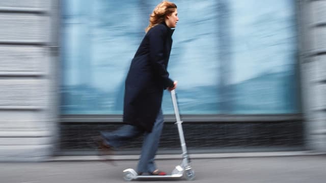 Eine Frau in Jeans und mit blauem Mantel flitzt mit einem Trottinett an einer Hausfassade vorbei. Eine Frau in Jeans und mit blauem Mantel flitzt mit einem Trottinett an einer Hausfassade vorbei.
