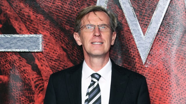 Ein Mann mit Brille und Anzug steht vor einer Wand, auf der Teile von riesigen Buchstaben zu erkennen sind. Ein Mann mit Brille und Anzug steht vor einer Wand, auf der Teile von riesigen Buchstaben zu erkennen sind.
