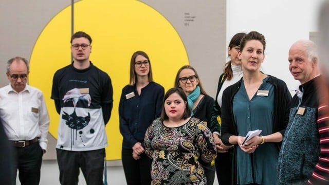 Eine Frau steht mit einer Gruppe von Menschen mit und ohne Behinderung vor einem abstrakten Gemälde. Eine Frau steht mit einer Gruppe von Menschen mit und ohne Behinderung vor einem abstrakten Gemälde.