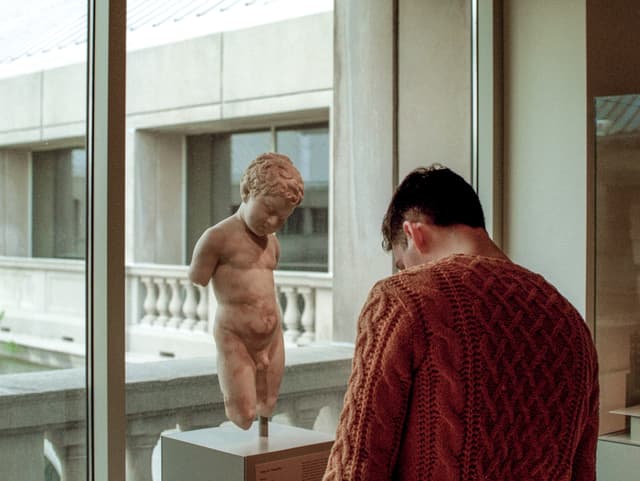 Ein Mann in orangem Pulli steht vor der Statue eines nackten Knaben und liest die Texttafel. Ein Mann in orangem Pulli steht vor der Statue eines nackten Knaben und liest die Texttafel.