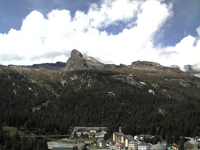Unten im Bild ist das Dorf zu sehen, in der Mitte ein Nadelwald mit einer markanten Felsnase über der Baumgrenze und darüber teils bewölkter, teils blauer Himmel.