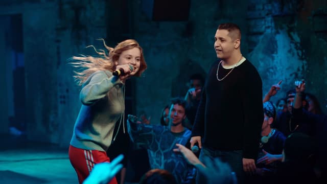 Auf der Bühne in einem Kellerclub: Ein Mädchen singt in ein Mikrofon, ein Mann steht daneben und schaut sie an. Im Hintergrund Menschen, die mit dem Handy filmen. Auf der Bühne in einem Kellerclub: Ein Mädchen singt in ein Mikrofon, ein Mann steht daneben und schaut sie an. Im Hintergrund Menschen, die mit dem Handy filmen.