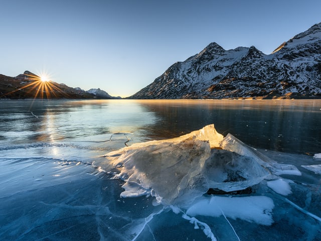 Gefrorener See, die Sonne hinter den Bergen knapp ersichtlich.