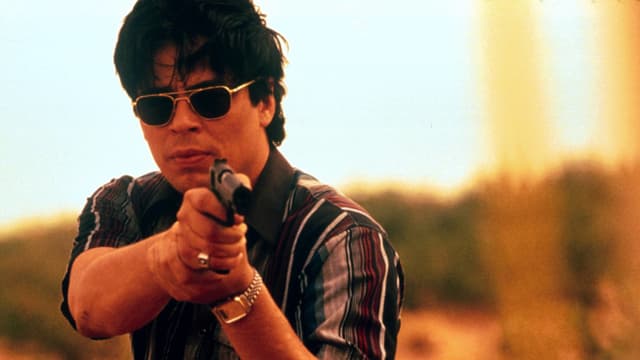 Bild aus dem Film "Traffic": Benicio del Toro steht in der Natur, trägt eine dunkle Sonnenbrille und zielt mit einer Pistole auf ein Tiel etwas unterhalb der Kamera. Bild aus dem Film "Traffic": Benicio del Toro steht in der Natur, trägt eine dunkle Sonnenbrille und zielt mit einer Pistole auf ein Tiel etwas unterhalb der Kamera.