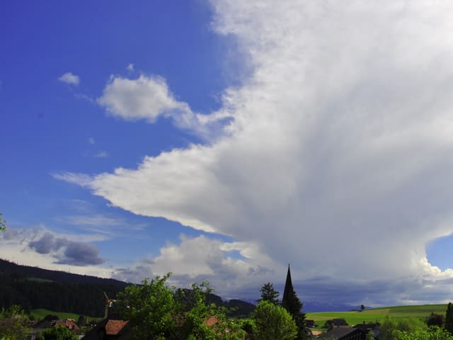 Links stahlblauer Himmel, rechts Wolkenfeld. Es kommt von Horizont her aus den Bergen. Am unteren Bildrand ist noch ein Dorf mit Wiesen zu sehen.