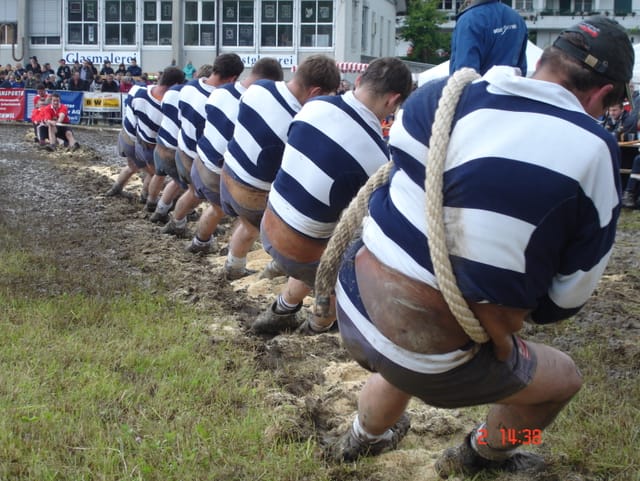 Mehrere Männer mit weiss-blau gestreiften Shirts stehen hintereinander und ziehen gemeinsam an einem Seil, an dessen Ende die Gegner Widerstand leisten.