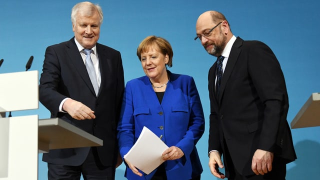 Drei Personen in einer Reihe vor einem Rednerpult stehend. Drei Personen in einer Reihe vor einem Rednerpult stehend.