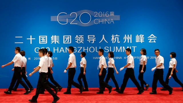 Chinesen gehen an einer blauen Wand vorbei mit grosser G20-Überschrift