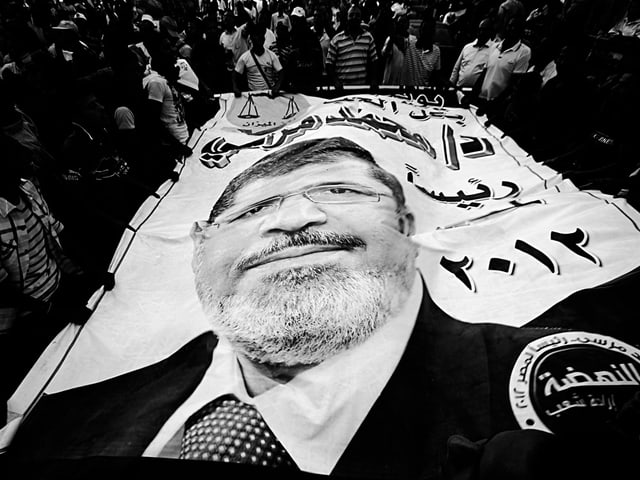 Schwarz-Weiss-Foto: Menschen halten eine grosse Fahne mit dem Bild von Mohammed Mursi. Durch die Unebenheiten der Fahne ist das Gesicht verzerrt. Schwarz-Weiss-Foto: Menschen halten eine grosse Fahne mit dem Bild von Mohammed Mursi. Durch die Unebenheiten der Fahne ist das Gesicht verzerrt.