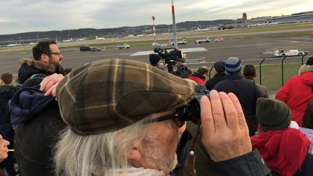 Mann mit grauen Haaren und Kappe sieht mit Feldstecher auf den Flughafen Mann mit grauen Haaren und Kappe sieht mit Feldstecher auf den Flughafen