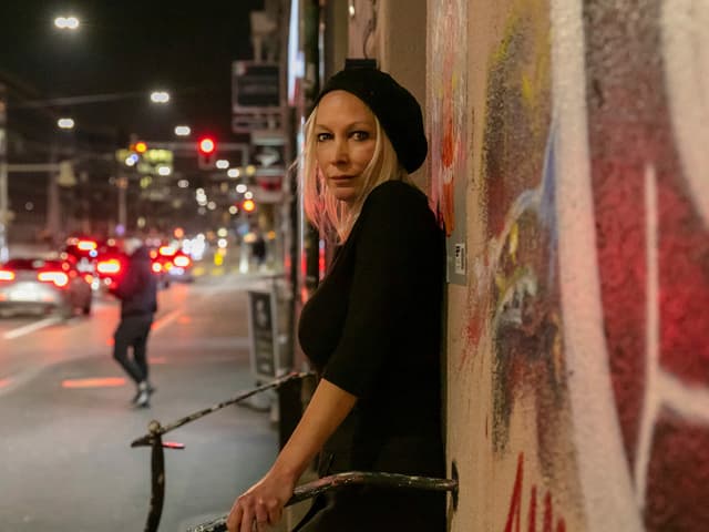 Frau mit blonden Haaren, schwarzer Wollmütze und schwarzem Shirt steht vor einem angesprayten Gebäude Frau mit blonden Haaren, schwarzer Wollmütze und schwarzem Shirt steht vor einem angesprayten Gebäude