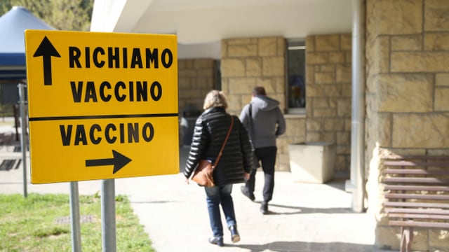Zwei Personen laufen zu einem Gebäude an einem Wegweiser Vaccino (Impfstoff) Zwei Personen laufen zu einem Gebäude an einem Wegweiser Vaccino (Impfstoff)