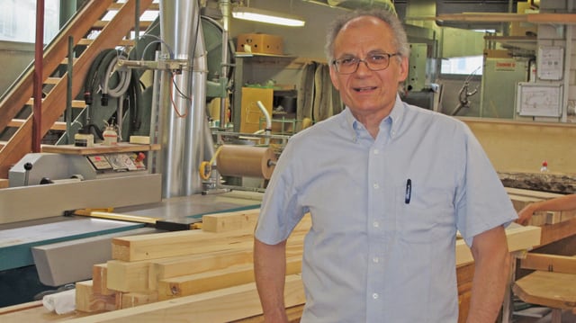 Ein Mann in blauem Hemd und Brille steht in einem Raum, umgeben von ganz viel Holz: Latten, Leiter und Werkzeug. Ein Mann in blauem Hemd und Brille steht in einem Raum, umgeben von ganz viel Holz: Latten, Leiter und Werkzeug.