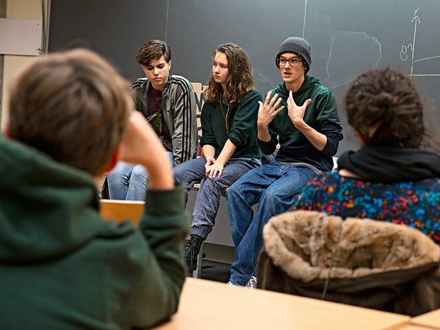 Drei Jugendliche sitzen auf dem Lehrerpult vor der Wandtafel und sprechen zu der Schulklasse. Drei Jugendliche sitzen auf dem Lehrerpult vor der Wandtafel und sprechen zu der Schulklasse.