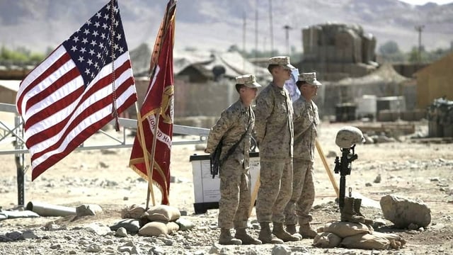 Soldaten stehen auf steinigem Boden, hintern ihnen weht eine US-Flagge