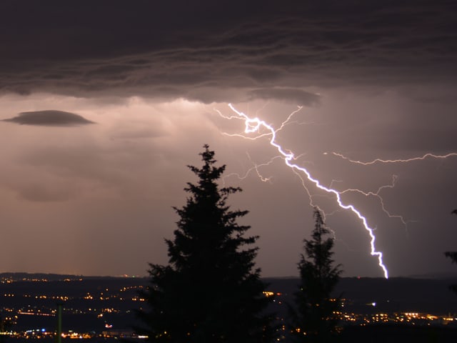 Im Vordergrund sind 2 Umrisse von Tannen zu sehen, im Hintergrund ein heller Blitz. Am Himmel hält sich eine Wolkendecke. Unten sind die Lichter der Siedlungen zu sehen.
